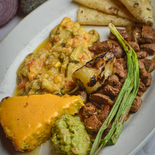 A Mexican dish with steak, roasted pepper, guacamole, cheesy corn casserole, and tortillas.
